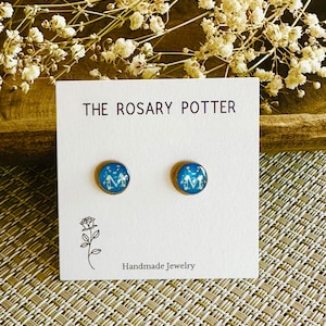 May include: A pair of blue and gold stud earrings with a white image of the Virgin Mary. The earrings are on a white card with the text "THE ROSARY POTTER" and "Handmade Jewelry".