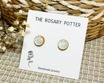 Puede incluir: Un par de pendientes de botón dorados con fondo blanco y una letra "V" azul rodeada de pequeñas flores. Los pendientes están sobre una tarjeta blanca con el texto "THE ROSARY POTTER Handmade Jewelry".