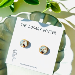 May include: A pair of gold stud earrings with a white background and a picture of an angel on each earring. The earrings are on a white card with the text "THE ROSARY POTTER" and "Handmade Jewelry".