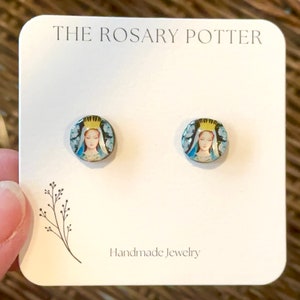May include: A pair of small, round, blue and gold earrings with a picture of the Virgin Mary on them. The earrings are on a white card with the text "THE ROSARY POTTER" and "Handmade Jewelry".