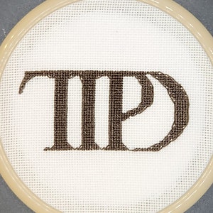 May include: A round, off-white cross-stitch embroidery hoop with the letters "TP" stitched in brown thread. The hoop has a light tan frame and a decorative metal loop at the top for hanging. The background is a solid gray.