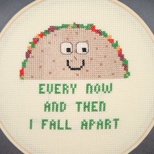May include: Cross-stitch embroidery in a wooden hoop features a smiling taco with the text "EVERY NOW AND THEN I FALL APART" in green. The taco is brown with colorful accents, and the background is off-white.