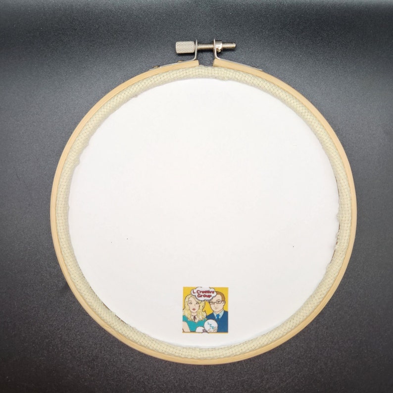May include: A round, wooden embroidery hoop with a white fabric center. The hoop is secured with a metal screw. A small, square graphic with two people and the text "L Creative Group" is at the bottom.