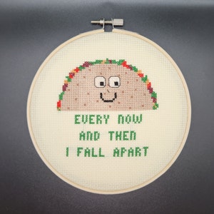 May include: Cross-stitch embroidery in a wooden hoop features a smiling taco with a brown shell, green, red, and yellow accents. The text "EVERY NOW AND THEN I FALL APART" is stitched below the taco in green.