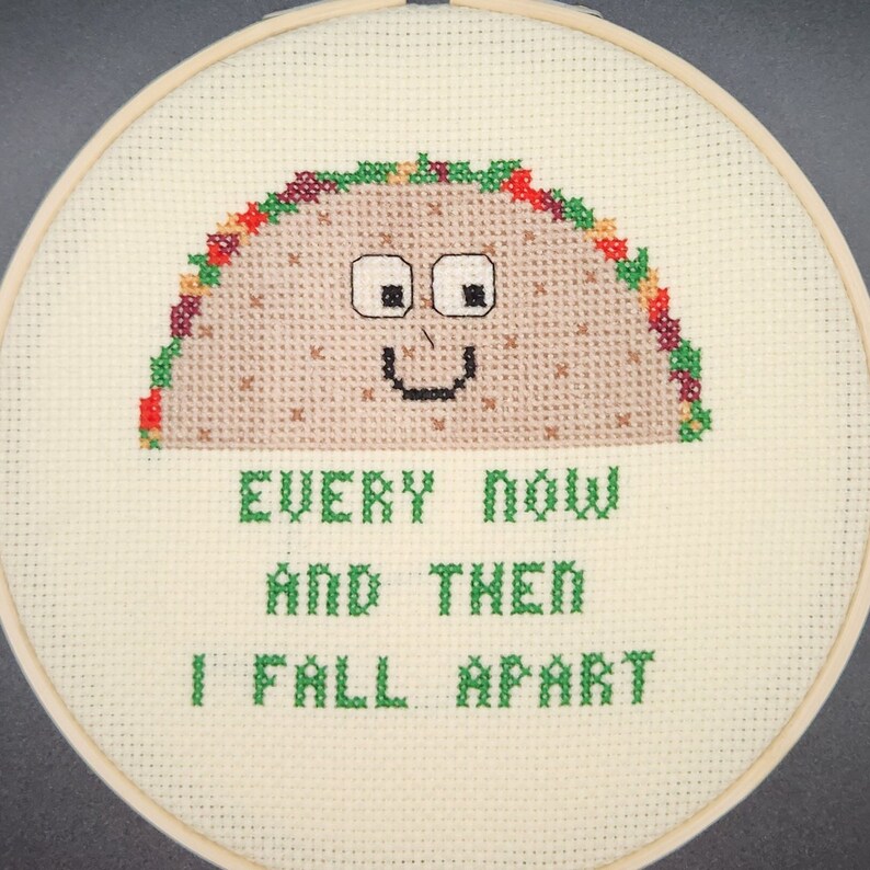 May include: Cross-stitch embroidery in a wooden hoop features a smiling taco with the text "EVERY NOW AND THEN I FALL APART" in green. The taco is brown with colorful accents, and the background is off-white.