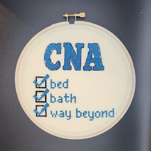 May include: A cross-stitch embroidery hoop with the word "CNA" in blue, above a checklist with "bed", "bath", and "way beyond" in blue. The embroidery is on a cream-colored fabric, and the hoop is made of wood.