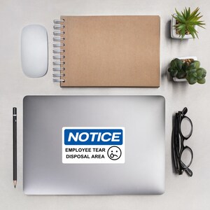 Op de afbeelding: Een blauwe en witte sticker met de tekst "NOTICE" in een blauw rechthoek. Onder de rechthoek staat de tekst "EMPLOYEE TEAR DISPOSAL AREA" in zwart met een verdrietige emoji.