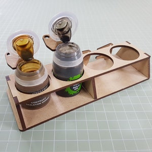 Puede incluir: Un soporte de madera para botes de pintura con tres ranuras. El soporte está hecho de madera marrón claro y tiene una etiqueta que dice "Contrast Skeleton Horde". Hay tres botes de pintura en el soporte, dos de los cuales están abiertos y vierten pintura marrón y negra.