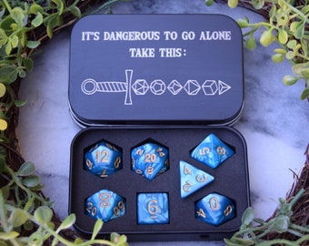 Può includere: Una scatola di metallo nero con il testo "It's dangerous to go alone. Take this:" e un'immagine di una spada. La scatola contiene un set di sette dadi blu e dorati.