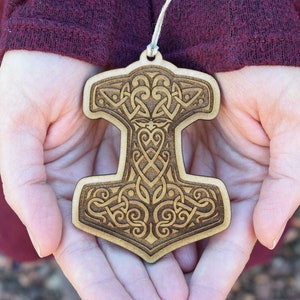 May include: A wooden ornament in the shape of Thor's hammer, featuring intricate Celtic knotwork. The light brown ornament is held in open hands, with a string for hanging. The background is blurred, with a maroon-colored sleeve visible.
