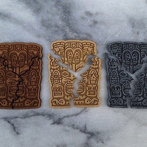 Puede incluir: Tres tallas de madera estilo tiki en tonos marr&oacute;n, beige y gris, cada una con un dise&ntilde;o agrietado. Las tallas presentan caras detalladas y patrones geom&eacute;tricos, sobre un fondo de m&aacute;rmol.