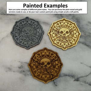 Puede incluir: Tres fichas circulares de madera con un dise&ntilde;o de calavera, cada una pintada de forma diferente. Una es gris, otra dorada y la otra marr&oacute;n c&aacute;lido. El texto "Painted Examples" y otro texto es visible en la parte superior de la imagen.