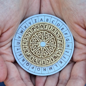 May include: A round, silver and wood cipher wheel with an outer ring of alphabet letters and an inner ring of symbols. The center features a detailed, carved wooden design. The wheel is held in open hands.