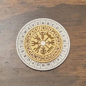 May include: A wooden, circular puzzle with a gold-colored center and a silver-colored outer ring. The center features intricate carvings and a central peg. The outer ring has letters and symbols. The puzzle is set on a dark wooden surface.