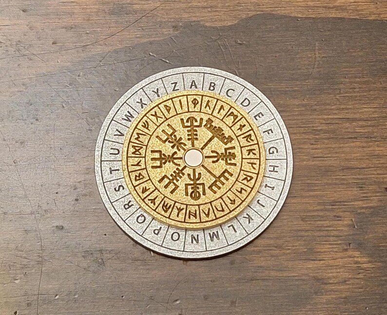 May include: A wooden, circular puzzle with a gold-colored center and a silver-colored outer ring. The center features intricate carvings and a central peg. The outer ring has letters and symbols. The puzzle is set on a dark wooden surface.