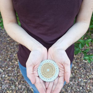 May include: A round, light blue and gold cipher disc with an alphabet around the edge and a maze-like design in the center. The disc is held in open hands, with a person wearing a maroon shirt and blue jeans in the background.