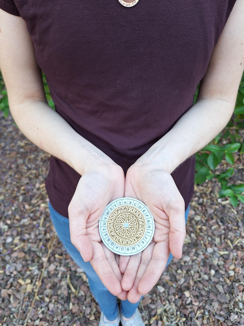 May include: A round, light blue and gold cipher disc with an alphabet around the edge and a maze-like design in the center. The disc is held in open hands, with a person wearing a maroon shirt and blue jeans in the background.