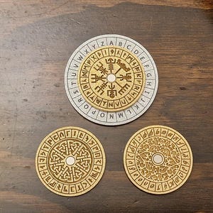 May include: Three wooden coasters with engraved designs. The top coaster is silver with an outer ring of letters and a central design. The other two coasters are gold-colored with intricate patterns. The coasters are on a dark wood surface.