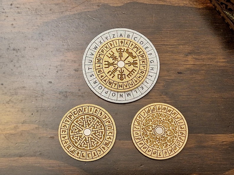 May include: Three wooden coasters with engraved designs. The top coaster is silver with an outer ring of letters and a central design. The other two coasters are gold-colored with intricate patterns. The coasters are on a dark wood surface.