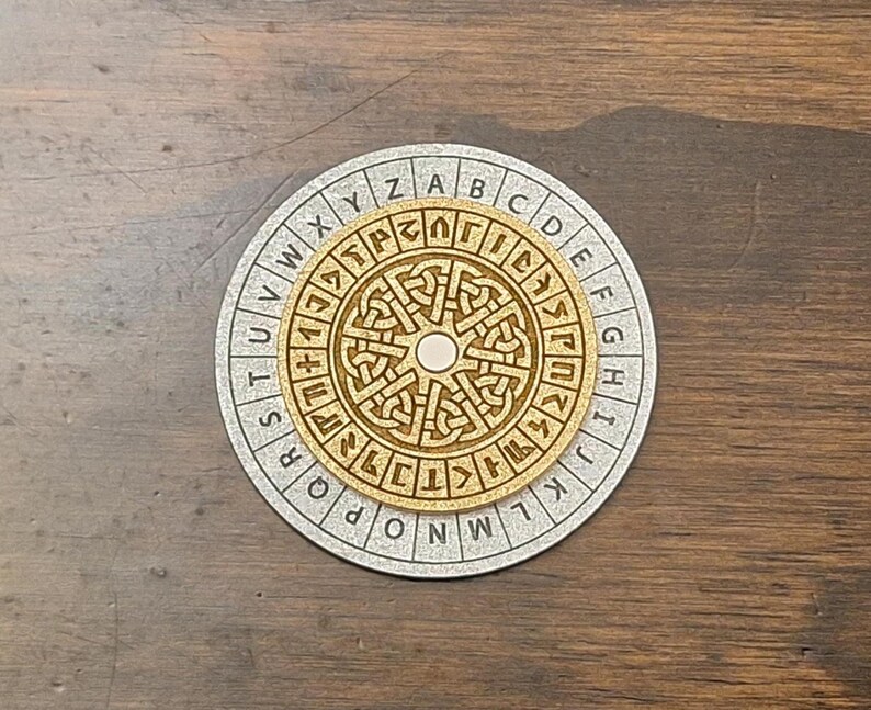 May include: A wooden cipher wheel with a circular design. The outer ring features the alphabet, while the inner circle displays intricate Celtic knot patterns. The wheel is set on a silver-colored base, creating a contrast with the natural wood tones.