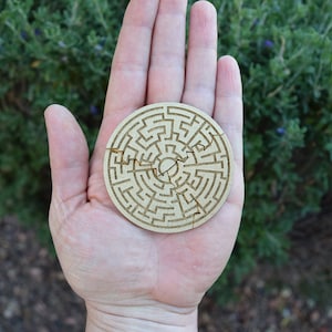 Puede incluir: Un rompecabezas redondo de madera con un dise&ntilde;o de laberinto. El rompecabezas, hecho de madera clara, se sostiene en una mano. El laberinto est&aacute; intrincadamente tallado en la madera, con varias piezas separadas.