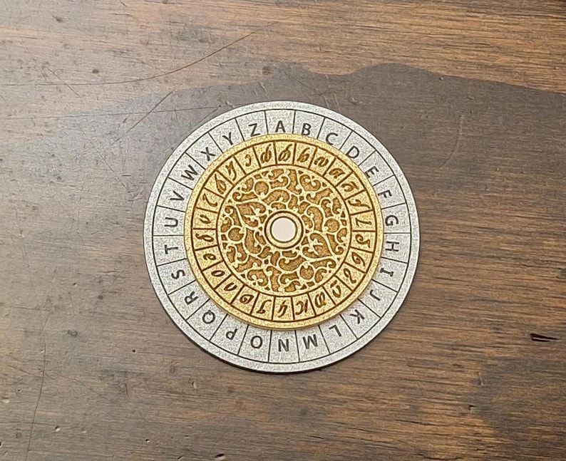May include: A wooden cipher wheel with a gold-colored center and a silver-colored outer ring. The center features intricate carvings and a central circle. The outer ring has the alphabet and a series of symbols. The wheel is designed for encoding and decoding messages.