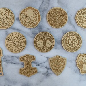 Puede incluir: Un conjunto de posavasos de madera cortados con l&aacute;ser con varios dise&ntilde;os, incluyendo un laberinto, una calavera, un pulpo y un escudo. Los posavasos son de color marr&oacute;n claro y est&aacute;n dispuestos sobre una superficie de m&aacute;rmol.