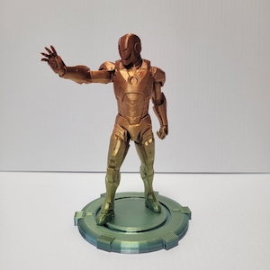 Puede incluir: Una figura de Iron Man impresa en 3D en un esquema de colores dorado y bronce, de pie sobre una base circular verde.