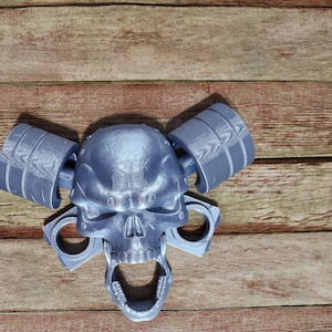 May include: A silver skull bottle opener with two silver cylinder-shaped handles on either side of the skull. The bottle opener is on a wooden surface.