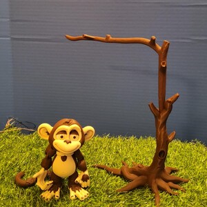 May include: A brown and cream-colored monkey figurine with large eyes and a smiling expression, positioned next to a brown, leafless tree branch structure with a wide, root-like base, both set against a plain blue background.