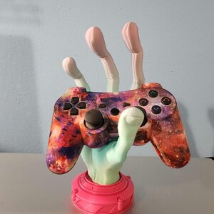 May include: A 3D-printed gaming controller stand shaped like a hand. The hand is a gradient of light green to pink, supporting a galaxy-themed controller. The base is bright pink.