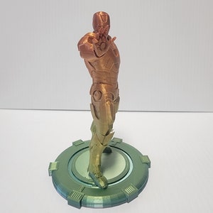 Puede incluir: Una figura de Iron Man impresa en 3D en un degradado de color dorado y bronce met&aacute;lico. La figura est&aacute; de pie sobre una base circular verde.