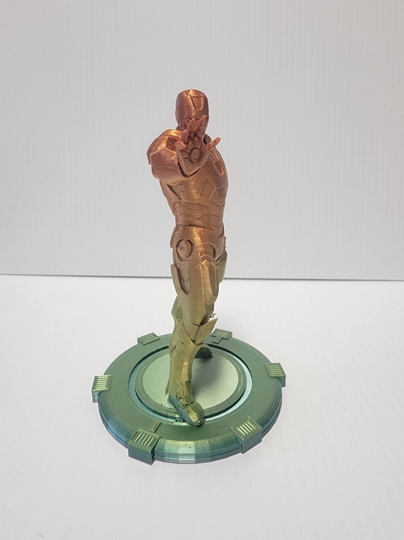Puede incluir: Una figura de Iron Man impresa en 3D en un degradado de color dorado y bronce met&aacute;lico. La figura est&aacute; de pie sobre una base circular verde.