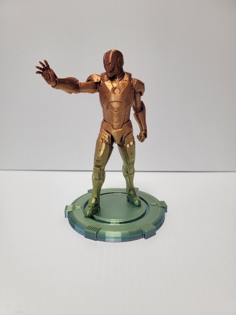Puede incluir: Una figura de Iron Man impresa en 3D en un esquema de colores dorado y bronce, de pie sobre una base circular verde.