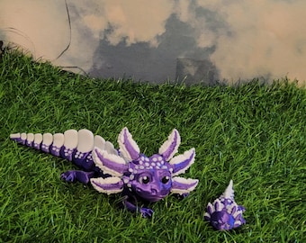 May include: Two purple and white axolotl figurines with white spines and spots. The larger axolotl is lying on its belly with its head turned to the side. The smaller axolotl is standing upright with its head tilted to the side. Both axolotls are on a green grassy surface.