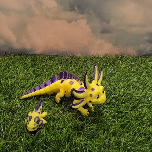 May include: Two yellow and purple axolotl figurines on a green grassy surface. The larger axolotl is standing on its hind legs and has a more detailed design. The smaller axolotl is lying on its belly.
