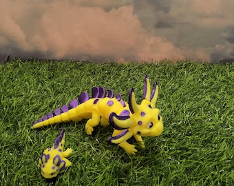 May include: Two yellow and purple axolotl figurines on a green grassy surface. The larger axolotl is standing on its hind legs and has a more detailed design. The smaller axolotl is lying on its belly.