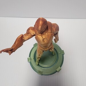 Puede incluir: Una figura de un superh&eacute;roe impresa en 3D de pie sobre una base circular verde. La figura es marr&oacute;n y tiene un dise&ntilde;o detallado.