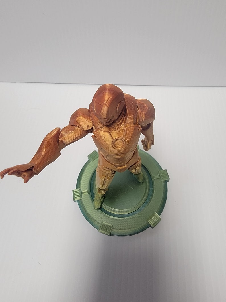 Puede incluir: Una figura de un superh&eacute;roe impresa en 3D de pie sobre una base circular verde. La figura es marr&oacute;n y tiene un dise&ntilde;o detallado.