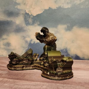 May include: A green and brown plastic figurine of a soldier kneeling and holding a weapon. The soldier is standing on a green and brown plastic base that resembles a destroyed building.