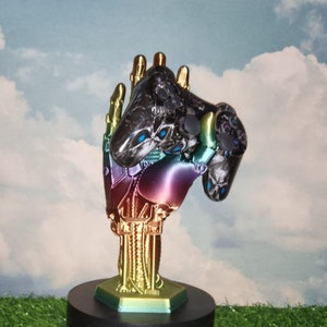 May include: A rainbow-colored robotic hand holding a black and green video game controller. The hand is on a black base with a hexagonal top.