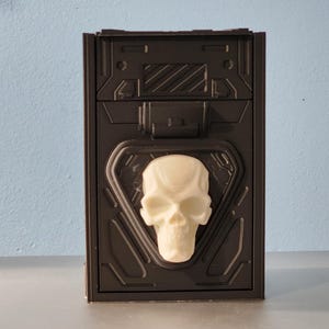 May include: A dark brown box with a white skull emblem. The box has a textured, geometric design and a hinged lid. The skull is centered on the front of the box.