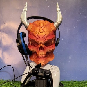 May include: A red and orange skull-shaped gaming headset stand with silver horns. The stand is holding a white wireless gaming controller with blue and black headphones.