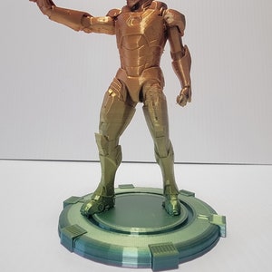 Puede incluir: Una figura de un superh&eacute;roe impresa en 3D con un acabado met&aacute;lico dorado y verde, de pie sobre una base circular verde.
