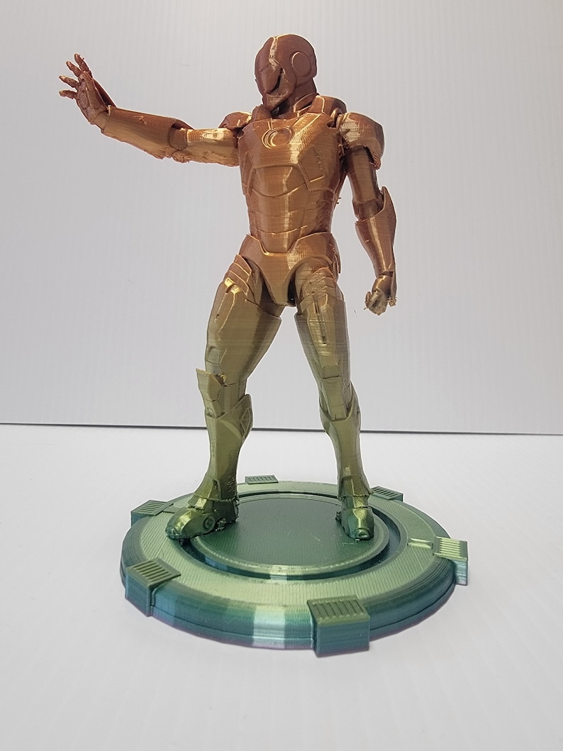Puede incluir: Una figura de un superh&eacute;roe impresa en 3D con un acabado met&aacute;lico dorado y verde, de pie sobre una base circular verde.