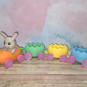 May include: A whimsical Easter train set featuring a gray bunny in an orange carrot car, followed by three pastel-colored egg-shaped cars. Each car has pink and purple wheels, creating a playful and festive scene.