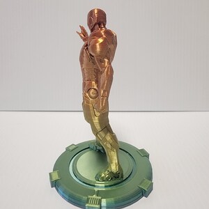 Puede incluir: Una figura de un superh&eacute;roe impresa en 3D de pie sobre una base circular. La figura est&aacute; hecha de un material met&aacute;lico y est&aacute; pintada en tonos dorados y cobrizos. La base est&aacute; pintada en tonos verdes.