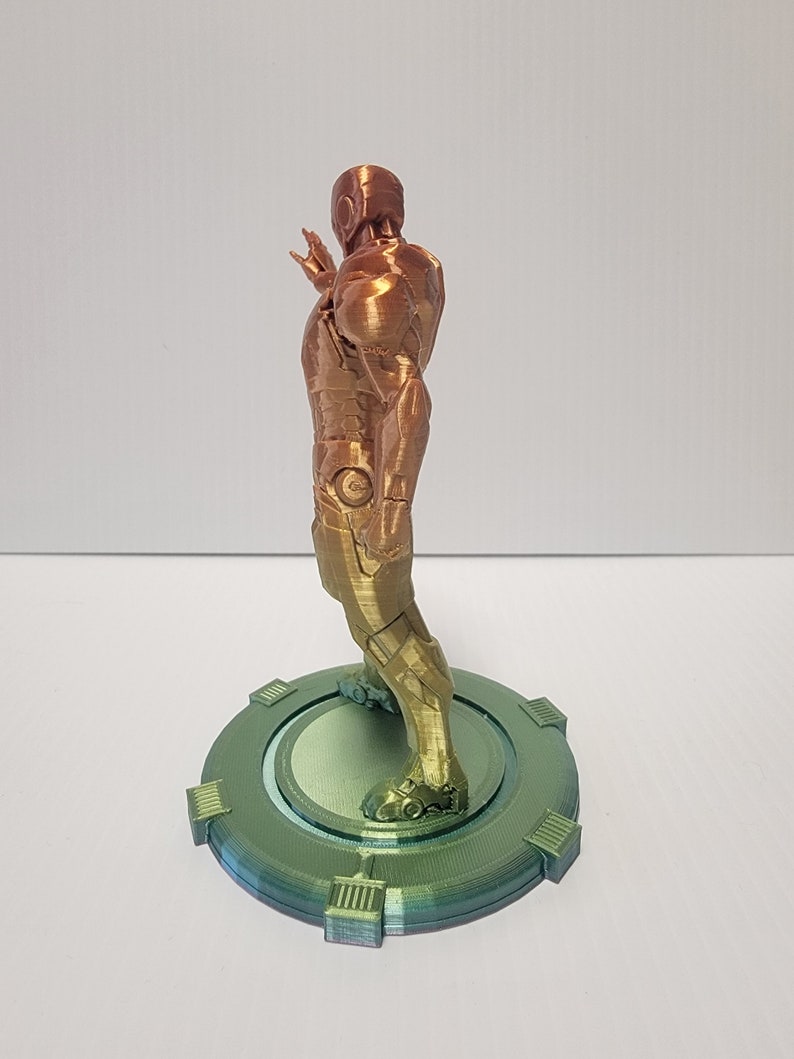 Puede incluir: Una figura de un superh&eacute;roe impresa en 3D de pie sobre una base circular. La figura est&aacute; hecha de un material met&aacute;lico y est&aacute; pintada en tonos dorados y cobrizos. La base est&aacute; pintada en tonos verdes.