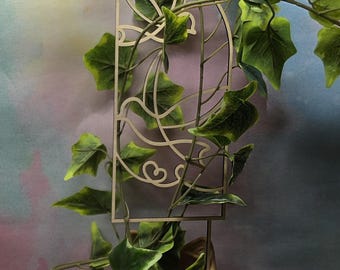 May include: A whimsical, beige-colored planter shaped like a smiling cat's face, holding green ivy. The ivy climbs a decorative, light-brown, arched trellis. The leaves are green with lighter green edges. The planter is on a dark surface.