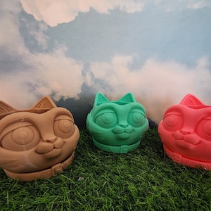 May include: Three ceramic cat planter pots in brown, green, and pink with large eyes and a collar. The pots are sitting on a green grassy surface.